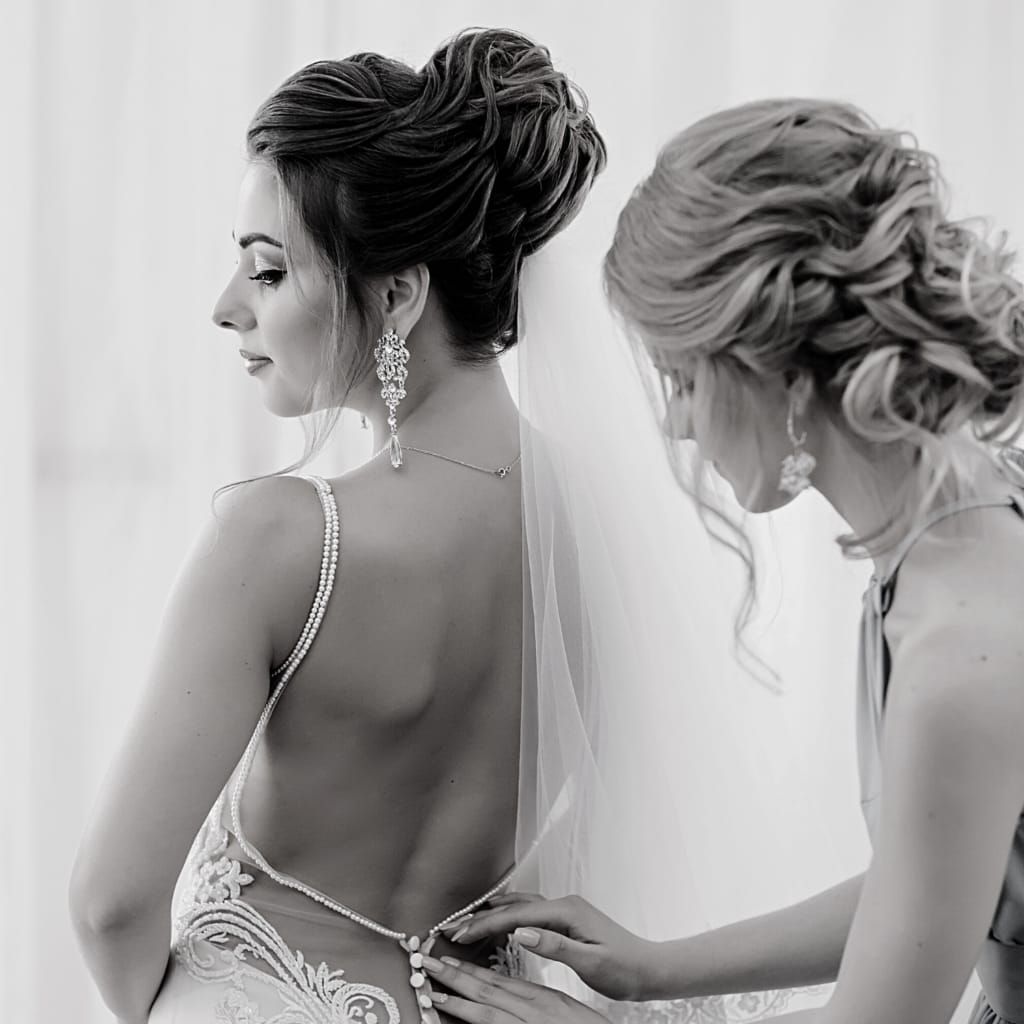 wedding hair updo view bride looking to side wearing open back wedding dress bridesmaid is buttoning wedding dress bridesmaids hair updo with curls