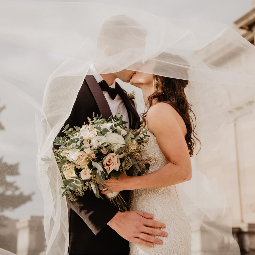 A bride and groom share a kiss under the bride's veil, showcasing the bride's beautifully styled bridal hair. She holds a bouquet of white and blush flowers with greenery and wears a lace wedding dress. The groom is in a black tuxedo, with the scene set outdoors in soft, natural light.