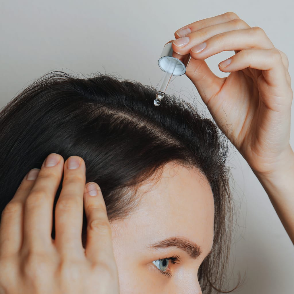 Person applying a few drops of almond oil to their scalp using a dropper, showcasing a hair care routine.