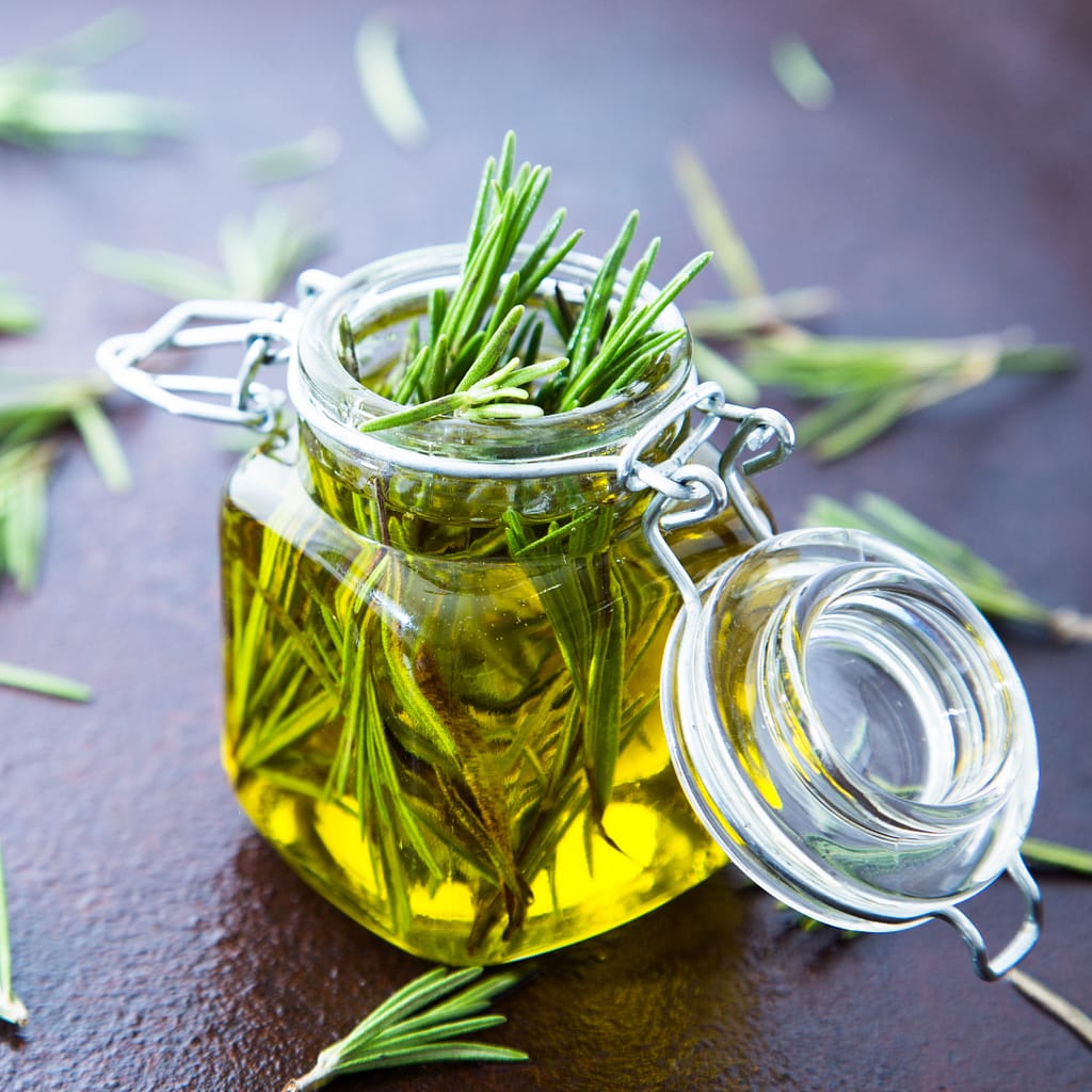 A small glass jar holds fresh rosemary sprigs submerged in warm water. This mixture, prepared for hair growth, will sit overnight to retain the natural oils and benefits of the herbs. Around the jar, rosemary sprigs are scattered on a dark surface, highlighting the natural ingredients used in this DIY rosemary water recipe for enhancing hair health.