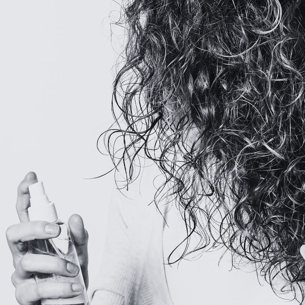 a woman has her head turned showcasing only her curly hair while she hold a spray bottle filled with rosemary water to help with hair growth