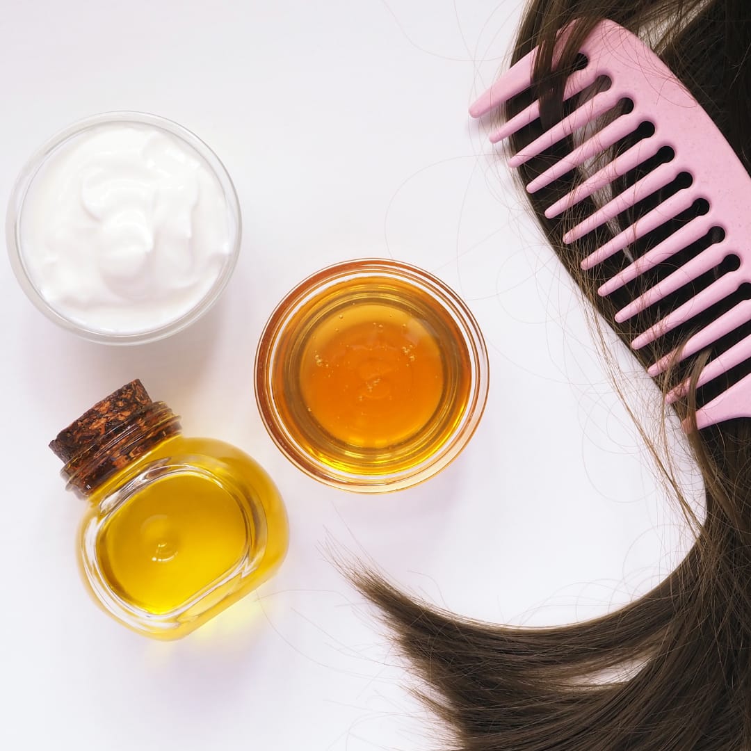 Top view of hair care products including a bottle of almond oil, a jar of cream, a small bowl of almond oil, and a pink wide-tooth comb, with strands of hair placed nearby.