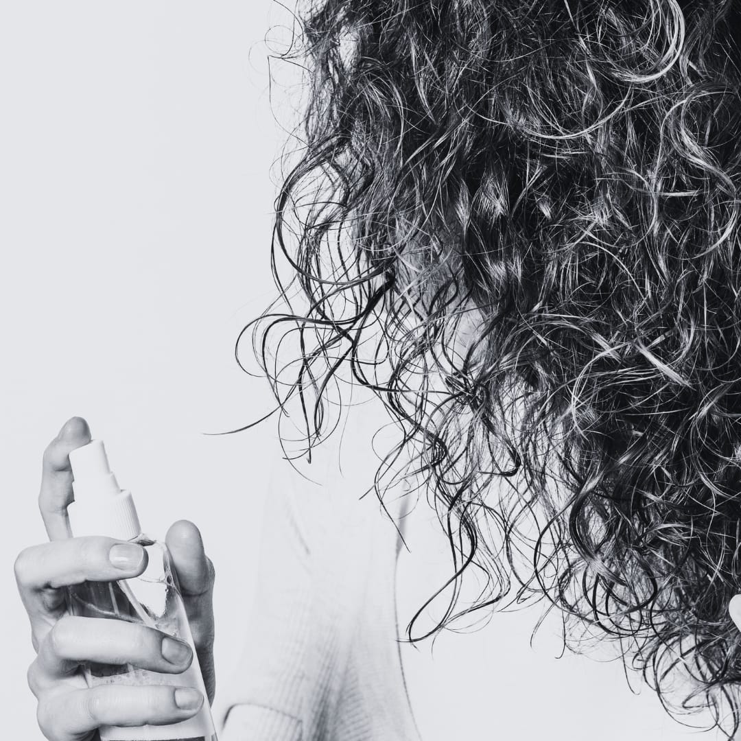 a woman has her head turned showcasing only her curly hair while she hold a spray bottle filled with rosemary water to help with hair growth