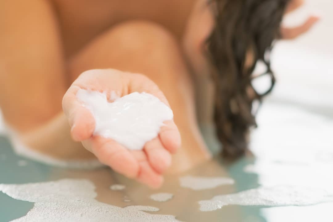 close up image of a woman's hand hold a hair mask solution sitting in a bathtub