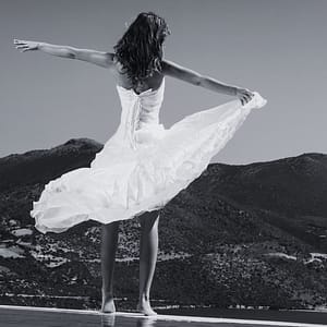 A woman in a white dress stands barefoot on a reflective surface, arms outstretched, with her back to the camera. She is stress free after using a bride guide. She faces a scenic mountainous landscape under a clear sky, the dress billowing in the breeze.