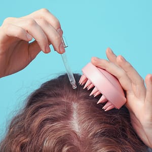 Person applying hair growth oils to the scalp with a dropper, using a scalp massager brush to stimulate hair follicles and enhance absorption.