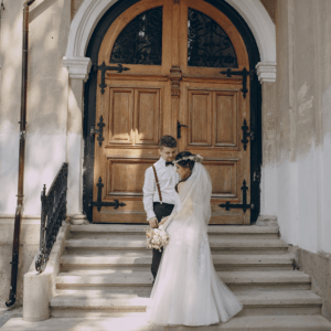 bridal hair updo curls bride wears a boho flower crown wedding hair accessory standing at wedding church stairs with groom wearing wedding dress and holding bridal flower bouquet