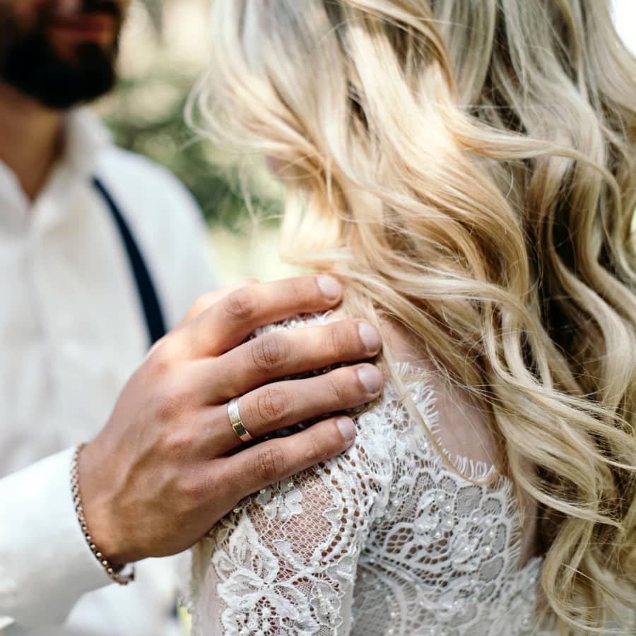 wedding hairstyle long hair curls close up view of bridal hairstyle with blonde hair highlights wearing lace wedding dress groom hand on her shoulder