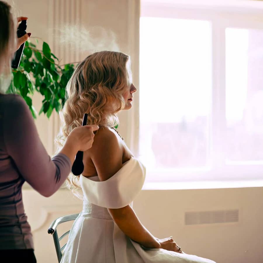 wedding hair bride glam waves mobile hairdresser spraying product to set bridal hairstyle for wedding day bride is wearing wedding dress